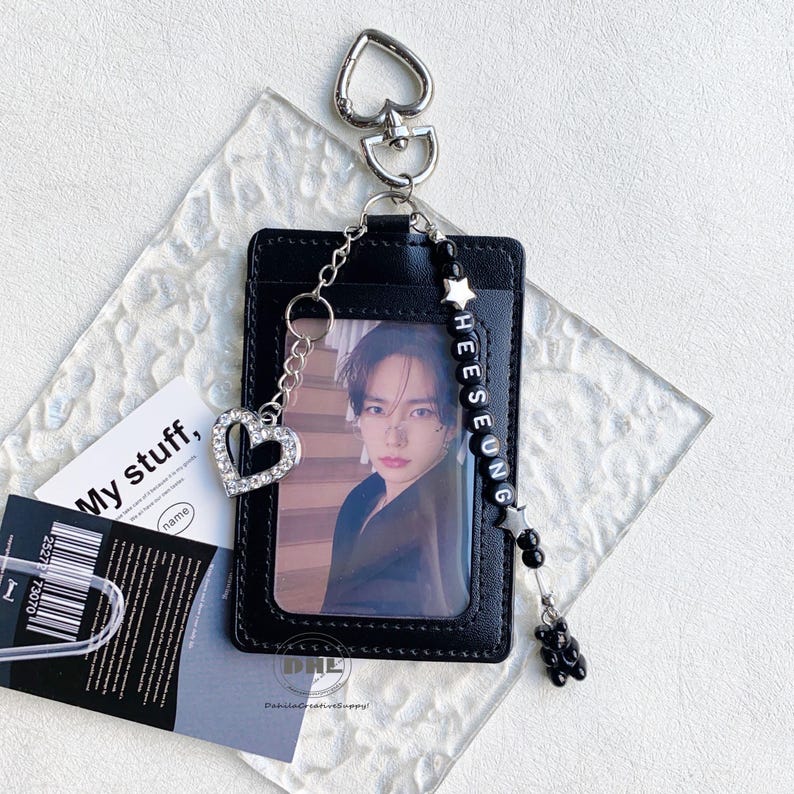 May include: Black cardholder with a photo, a silver heart-shaped charm, and a beaded charm with the name "HEESEUNG". The cardholder has a silver heart-shaped keychain. A small black bear charm hangs from the beaded charm.