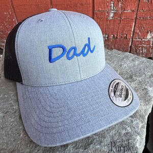 May include: A light grey trucker cap with a black mesh back. The word "Dad" is embroidered in blue on the front. The cap has a curved peak and a snapback closure. The cap is sitting on a grey rock.