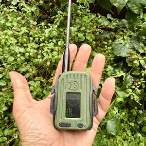 May include: A green and black handheld radio with a black antenna. The radio has a small screen displaying text and a compass. The radio is held in a hand.