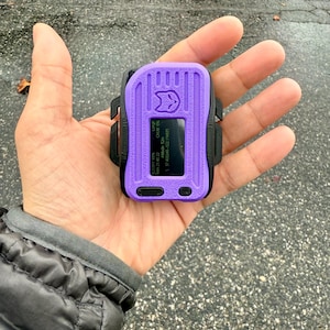 Könnte beinhalten: Ein kleines, violettes Handgerät mit einem schwarzen Band und einem Bildschirm, der Text und Zahlen anzeigt. Das Gerät hat ein Katzen-Symbol auf der Vorderseite.