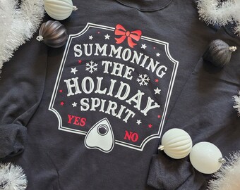 Op de afbeelding: Zwart sweatshirt met de tekst "SUMMONING THE HOLIDAY SPIRIT" in wit en rood. Het ontwerp bevat een strik, sneeuwvlokken, sterren en een planchette. Witte en donkergrijze ballen omringen het sweatshirt.