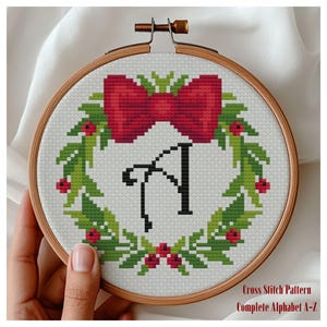 May include: A circular wooden embroidery hoop containing a cross-stitch pattern of a green holly wreath with red berries and a large red bow at the top. Centered within the wreath is the black letter A. The text Cross Stitch Pattern Complete Alphabet A-Z is visible.
