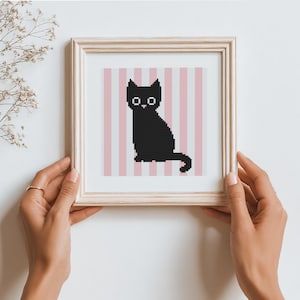 May include: A framed cross-stitch art piece featuring a black cat silhouette against a pink and white striped background. The cat has simple, geometric features. The frame is a light wood color.