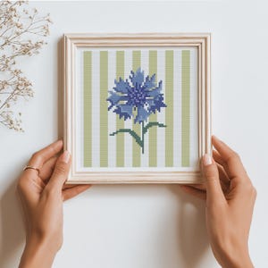 May include: A framed cross-stitch artwork featuring a blue flower with green leaves and stem against a striped background of white and light green. The artwork is in a light wood frame.