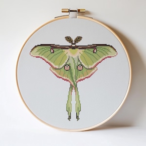 May include: A cross-stitch embroidery of a Luna moth, displayed in a wooden hoop. The moth features a detailed design with shades of green, pink, and brown. The wings are spread, showcasing the intricate pixelated pattern. The hoop is approximately 8 inches in diameter.