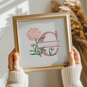 May include: A square gold frame containing a cross-stitch embroidery on white fabric. The design features a large pink letter C with the name Chandler stitched in black across the center, accompanied by pink carnation flowers and green stems.