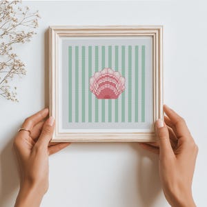 May include: A square wooden frame containing a cross-stitch embroidery piece. The design features a pink seashell centred against a background of vertical light green and white stripes. The embroidery is displayed within a light-coloured wood frame.