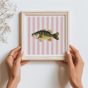 May include: A square framed cross-stitch artwork featuring a green and brown fish centered against a background of vertical light pink and white stripes. The piece is enclosed in a light-colored wooden frame with a simple, clean profile.