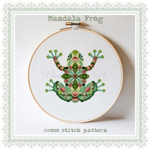 May include: A cross stitch pattern of a green frog with a colorful mandala design on its back. The pattern is displayed in a wooden embroidery hoop. The text "Mandala Frog" and "cross stitch pattern" are below the hoop.