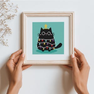 May include: Framed cross-stitch art featuring a black cat decorated as a Christmas tree. The cat has a gold star on its head and is wrapped in colorful Christmas lights. The background is a teal color.