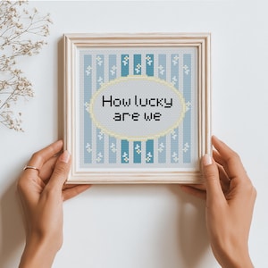 May include: A square cross-stitch embroidery in a light wood frame featuring vertical blue and white stripes with small white flower accents. A central yellow oval contains the black text How lucky are we. The design is centered within the frame.