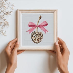 May include: A square framed cross-stitch embroidery featuring a heart-shaped cherry with a brown and tan leopard print pattern. The cherry is topped with a light brown stem and a large, bright pink bow tied in a decorative knot at the center.