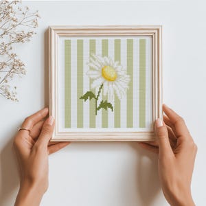 May include: A framed cross-stitch artwork featuring a white daisy with a yellow center and green leaves. The background has vertical stripes in light green and white. The frame is light wood.