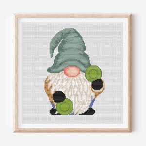 May include: Cross-stitch art of a gnome holding green coins. The gnome has a long white beard, a pink nose, and a green hat. The artwork is framed in a light brown wooden frame and set against a white background.