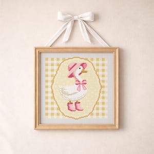 May include: A framed cross-stitch depicting a white goose adorned with a pink hat, bow, and boots. The goose is set against a yellow oval on a yellow and white gingham backdrop. The frame is light brown, and the artwork is hung by a white ribbon.