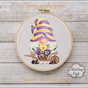 May include: A cross-stitch pattern of a purple and yellow gnome holding a wheelbarrow full of flowers. The gnome is wearing a yellow hat with purple stripes and has a white beard. The wheelbarrow is brown and has a black handle. The flowers are purple, yellow, and white. The background is a light gray fabric.