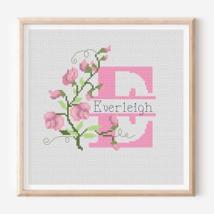 May include: Cross-stitch artwork in a wooden frame. A large pink letter "E" is the focal point, with the name "Everleigh" in white. Pink flowers and green leaves decorate the left side of the letter. The background is a light grey.