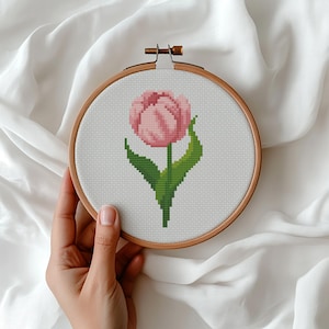 Könnte beinhalten: Ein Kreuzstich-Stickrahmen mit einer rosa Tulpe mit grünen Blättern und Stiel. Das florale Design befindet sich vor einem weißen Hintergrund, der von einem Holzrahmen umgeben ist. Das Bastelprojekt wird vor weißem Stoff gehalten.