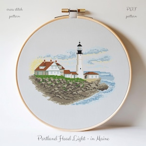 May include: A cross stitch pattern in a wooden embroidery hoop featuring the Portland Head Light in Maine. The design depicts a white lighthouse and buildings on a rocky shore with blue water and a pale yellow sun. Text reads cross stitch pattern, PDF pattern, and Portland Head Light - in Maine.
