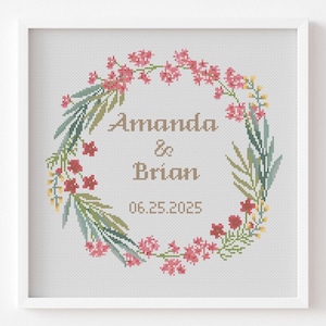 May include: A framed cross-stitch featuring a floral wreath design. The wreath is composed of pink flowers, green leaves, and yellow accents. The names "Amanda & Brian" and the date "06.25.2025" are stitched in the center.