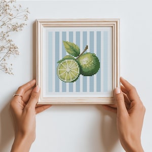 May include: Cross-stitch artwork of green limes, one sliced, with leaves, set against a light blue and white striped background. The artwork is framed in a light wood frame. The overall theme is a refreshing, summery fruit design.
