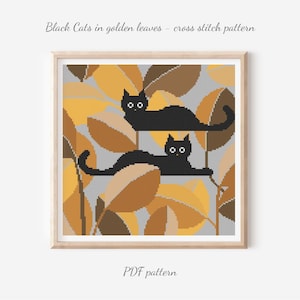 Pode incluir: Padrão de ponto cruz com dois gatos pretos entre folhas douradas, sobre um fundo cinzento. A obra está emoldurada em madeira clara. O texto diz "Black Cats in golden leaves - cross stitch pattern" e "PDF pattern".