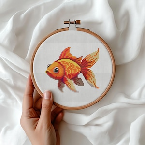 Op de afbeelding: Kruissteekborduurwerk van een goudvis in een houten hoepel. De vis is voornamelijk oranje en geel met rode accenten, en de hoepel wordt op zijn plaats gehouden met een messing schroef. De achtergrond is wit.