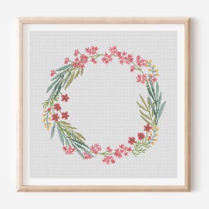 May include: A framed cross-stitch floral wreath. The wreath features pink flowers, green leaves, and yellow accents, arranged in a circular pattern. The artwork is set against a white background and framed in a light wood.