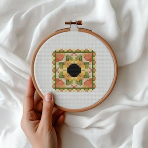 May include: A cross-stitch embroidery hoop with a sunflower design. The design features a black centre, surrounded by yellow, green, and orange details. The embroidery is set in a wooden hoop, held against a white fabric background.