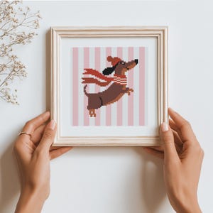 Pode incluir: Uma obra de arte em ponto cruz emoldurada com um dachshund marrom usando um cachecol e chapéu vermelhos, em um fundo listrado rosa e branco. O dachshund parece estar pulando. A moldura é de madeira clara.