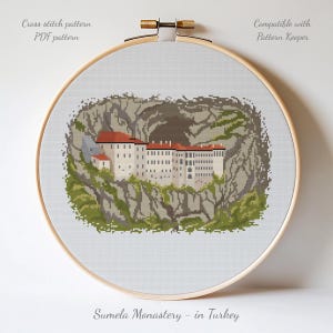 May include: A cross stitch pattern in a wooden embroidery hoop featuring the Sumela Monastery in Turkey. The design depicts a white building with a red roof nestled into a rocky cliffside with green foliage. Text reads Cross stitch pattern, PDF pattern, Compatible with Pattern Keeper, and Sumela Monastery - in Turkey.