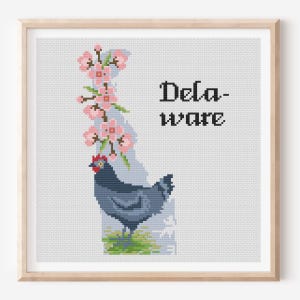 May include: A framed cross-stitch pattern featuring the state of Delaware in light blue, decorated with pink cherry blossoms and a dark blue chicken with a red comb. The text Delaware is stitched in black, set against a white grid background.