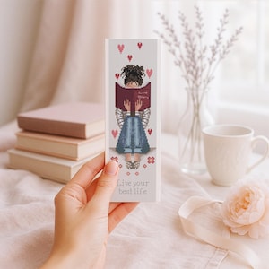 May include: A bookmark featuring a cross-stitch design of a person reading a book with the words "Love Story" visible. The bookmark has a white background with red hearts and the text "live your best life".