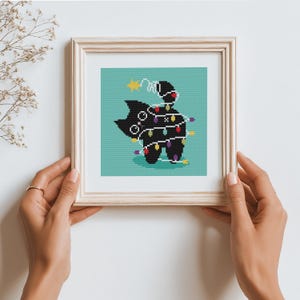 May include: Framed cross-stitch art depicting a black cat in a cape, adorned with colourful Christmas lights, set against a teal backdrop. A gold star is positioned above the cat. The frame is a light wood colour.