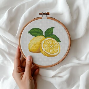 Puede incluir: Un bastidor de bordado circular de madera que contiene un patrón de punto de cruz de un limón amarillo entero y una mitad de limón cortada con hojas verdes. El diseño está bordado sobre tela blanca, centrado dentro del marco.
