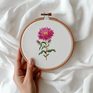 May include: A cross-stitch embroidery of a pink aster flower with a yellow center, green stem, and leaves, set in a wooden embroidery hoop. The floral design is on white fabric.