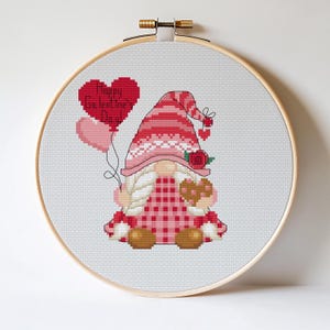 Puede incluir: Bordado de punto de cruz en un aro de madera que presenta un gnomo de San Valentín sosteniendo globos en forma de corazón y un dulce en forma de corazón. El gnomo lleva un sombrero rosa y rojo y un atuendo a cuadros. El texto "Happy Galentine's Day!" está en un globo en forma de corazón.