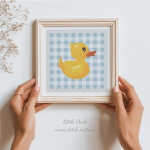 Könnte beinhalten: Ein gerahmtes Kreuzstichmuster mit einer gelben Ente mit orangefarbenem Schnabel und schwarzem Auge, vor einem blau-weiß karierten Vichy-Hintergrund. Der Rahmen ist aus hellem Holz. Der Text "Little Duck cross stitch pattern" steht darunter.