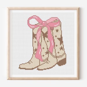 Pode incluir: Uma obra de arte em ponto cruz emoldurada com uma bota de cowboy bege com padrões de estrelas e cruzes castanhas. Um grande laço rosa adorna a parte superior da bota. A obra de arte é apresentada numa moldura de madeira castanha clara.