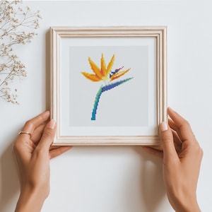 May include: A framed cross-stitch art piece featuring a vibrant Bird of Paradise flower. The flower has orange and yellow petals, a blue and purple beak, and a green and blue stem. The artwork is displayed in a light-colored wooden frame.