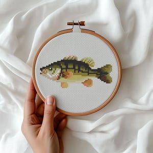 May include: A circular wooden embroidery hoop containing a cross-stitch pattern of a fish. The fish is rendered in shades of dark green, olive, and light yellow, featuring a detailed scale pattern and fins, centered on a white fabric background.