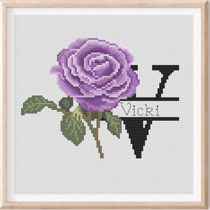 Op de afbeelding: Kruissteekkunst met een grote paarse roos met groene bladeren en een bruine stengel. Een zwarte letter "V" staat naast de roos, met de naam "Vicki" in witte tekst. Het kunstwerk zit in een houten lijst.