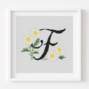 Può includere: Opera d'arte a punto croce incorniciata con la lettera "F" in nero, circondata da margherite bianche con centri gialli e foglie verdi. L'opera è ambientata su uno sfondo grigio chiaro e incorniciata in una cornice quadrata bianca.
