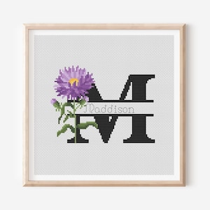 以下が含まれることがあります： 黒い文字「M」と筆記体で「Maddison」という名前が刺繍されたクロスステッチアート。 黄色の中心と緑の葉を持つ紫色の花が文字の左側に配置されています。 作品は木製のフレームに入っています。
