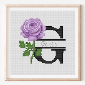 May include: Cross-stitch artwork showing a purple rose and the letter "G" in black. The name "Gayla" is written in cursive within the letter. The artwork is framed in a light wood frame.