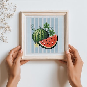 Puede incluir: Obra de arte de punto de cruz de una sandía, con una fruta entera y una rodaja. El diseño está sobre un fondo a rayas azul claro, enmarcado en madera clara. La sandía es verde con rayas oscuras, y la rodaja es roja con semillas negras.