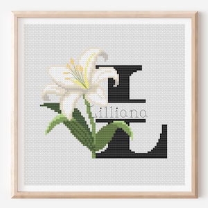 以下が含まれることがあります： 白いユリと黒い文字「E」を特徴とするクロスステッチアート。下には「Lilliana」という名前があります。このアートワークは、白い背景に対して、明るい木製のフレームに収められています。