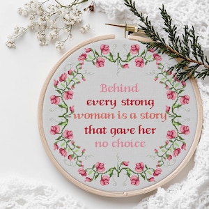 Könnte beinhalten: Ein Kreuzstich-Stickrahmen mit dem Zitat "Hinter jeder starken Frau steckt eine Geschichte, die ihr keine Wahl ließ" in rosa Schrift. Das Design ist von einem floralen Rand aus rosa Blumen und grünen Ranken umgeben.
