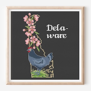 May include: A framed cross-stitch embroidery on a dark gray background featuring a blue chicken standing on a green patch, a vertical branch with pink peach blossoms, and the white text Delaware in a pixelated font style within a light wood frame.