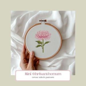 May include: A cross-stitch embroidery hoop featuring a pink chrysanthemum flower. The flower is stitched on white fabric with green stem and leaves. The text "Mini Chrysanthemum cross stitch pattern" is below the hoop.